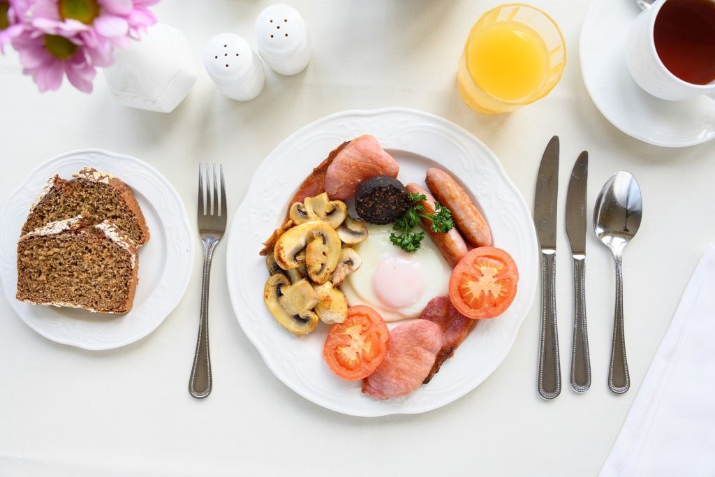Een bord met een warm ontbijt: gebakken ei, spek, worst, bloedworst, gegrilde tomaat, champignons en peterselie. Ernaast liggen bruin brood met boter, een glas sinaasappelsap, een kop thee en bestek op een wit tafelkleed. Roze bloemen linksboven. | Echt Ierland Vakanties