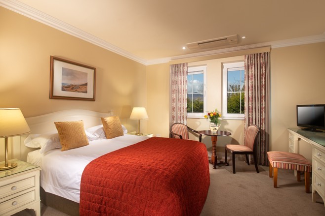 Een gezellige hotelkamer met een tweepersoonsbed met een rood dekbed, twee bruine kussens, nachtkastjes met lampen, een klein rond tafeltje met bloemen en twee gestreepte stoelen, een tv op een bureau en twee ramen met gordijnen waardoor daglicht binnenkomt. | Echt Ierland Vakanties