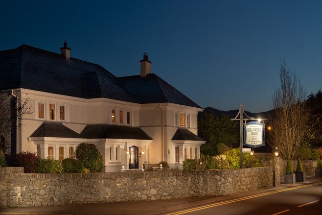Een witte lodge van twee verdiepingen met gloeiende ramen staat achter een stenen muur in de schemering. Het gebouw heeft een donker, steil dak met schoorstenen. Een verlicht bord bij de ingang vermeldt Killarney Lodge. Bomen en struiken omringen het terrein en de lucht is diepblauw. | Echt Ierland Vakanties