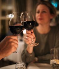 Een man en een vrouw zitten aan een restauranttafel en proosten met glazen rode wijn. De vrouw is enigszins onscherp en lacht, terwijl zacht kaarslicht en een elegant decor een warme, intieme sfeer creëren. Andere tafels zijn zichtbaar in de wazige achtergrond. | Echt Ierland Vakanties