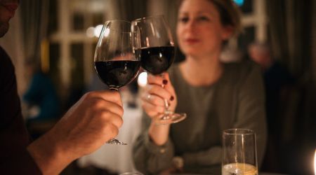 Een man en een vrouw zitten aan een restauranttafel en proosten met glazen rode wijn. De vrouw is enigszins onscherp en lacht, terwijl zacht kaarslicht en een elegant decor een warme, intieme sfeer creëren. Andere tafels zijn zichtbaar in de wazige achtergrond. | Echt Ierland Vakanties