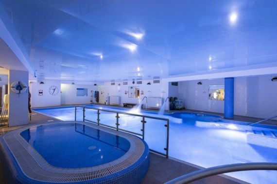 Overdekt zwembad met felblauwe verlichting. De ruimte omvat een ronde hottub op de voorgrond, gescheiden door een reling van een groter zwembad op de achtergrond. De zwembadelementen omvatten fonteinen, een klok, witte muren en zwembadapparatuur. | Echt Ierland Vakanties