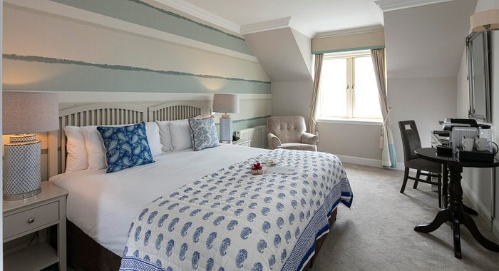 Een lichte, netjes ingerichte hotelkamer met een groot bed met een wit-blauwe deken, kussens met blauwe dessins en nachtkastjes met lampen. Er is een raam met lichtgordijnen, een bureau met een stoel en een theeservies op een ronde tafel. | Echt Ierland Vakanties