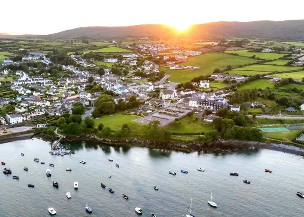 Luchtfoto van een kustdorpje bij zonsondergang, met kleine bootjes voor anker in kalm water. Het stadje heeft witte huizen en groene velden, omringd door glooiende heuvels. De zon zakt achter de heuvels en werpt een gouden gloed over het tafereel. | Echt Ierland Vakanties