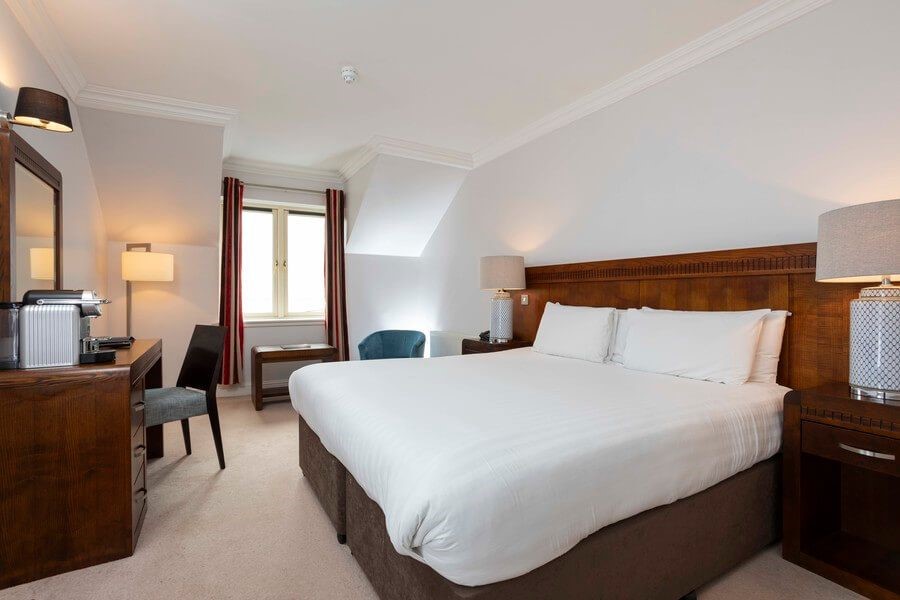 Een netjes opgemaakt tweepersoonsbed met wit linnen staat in een lichte hotelkamer. De kamer is voorzien van houten meubels, twee bedlampjes, een bureau met stoel, een blauwe fauteuil en een raam met rode gordijnen die natuurlijk licht binnenlaten. | Echt Ierland Vakanties