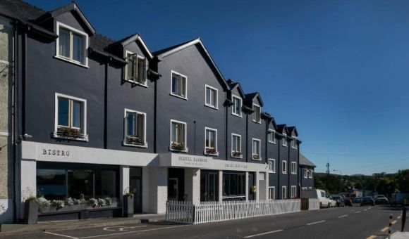 Een rij moderne, donkergrijze gebouwen met witte accenten staat aan een zonnige straat. De winkelpuien op de begane grond omvatten een bistro en wat lijkt op een hotelingang, terwijl de bovenste verdiepingen witte kozijnen en bloembakken hebben. De lucht is helderblauw. | Echt Ierland Vakanties