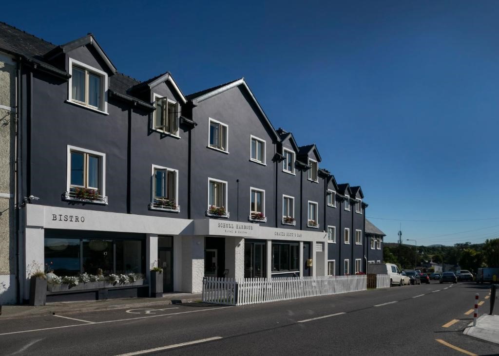 Een rij moderne, donkergrijze gebouwen met witte accenten staat aan een zonnige straat. De winkelpuien op de begane grond omvatten een bistro en wat lijkt op een hotelingang, terwijl de bovenste verdiepingen witte kozijnen en bloembakken hebben. De lucht is helderblauw. | Echt Ierland Vakanties