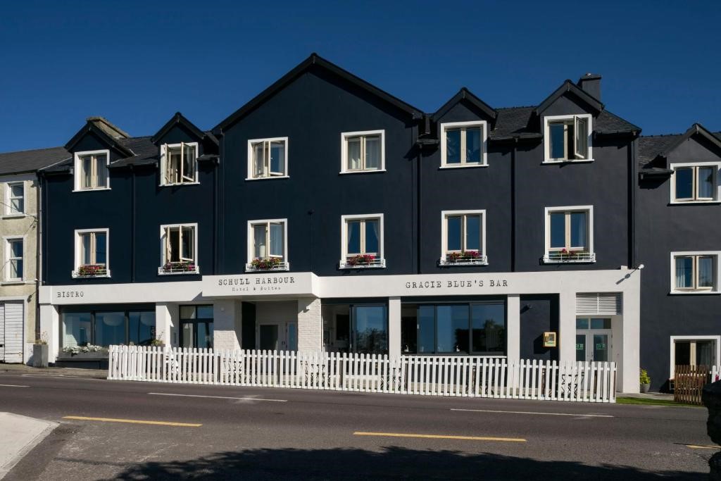 Een modern, donkerblauw gebouw van drie verdiepingen met witte accenten, grote ramen en bloembakken met bloemen. Boven de ingangen hangen de borden Schull Harbour en Gracie Blue's Bar. Een wit hek langs de stoep; de lucht is helderblauw. | Echt Ierland Vakanties