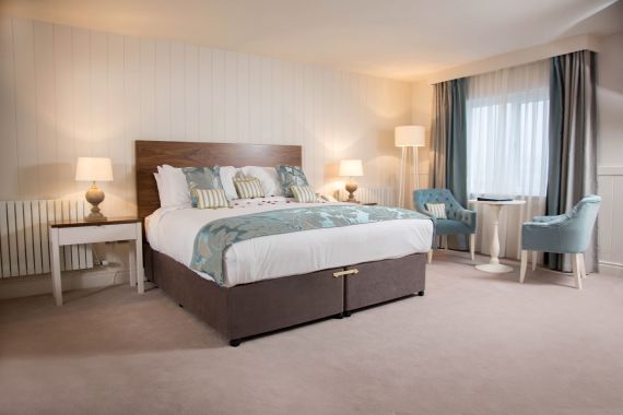 Een gezellige slaapkamer met een groot bed met een houten hoofdeinde en een blauw bloemensprei. Twee nachtkastjes met lampen flankeren het bed. Een raam met doorschijnende gordijnen en draperieën laat natuurlijk licht binnen. Twee blauwe stoelen en een kleine ronde tafel staan bij het raam. De kamer heeft beige tapijt. | Echt Ierland Vakanties