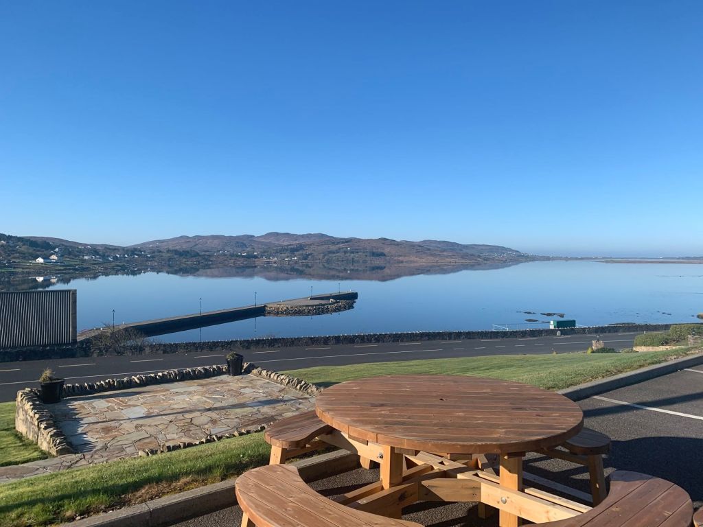 Een houten picknicktafel kijkt uit over een kalm water met een helderblauwe lucht. Op de achtergrond rijzen heuvels zachtjes op langs de horizon. Een stenen platform en goed onderhouden gras leiden naar een weg, waarachter een kalme baai de lucht weerspiegelt. Een verre pier strekt zich uit in het water. | Echt Ierland Vakanties