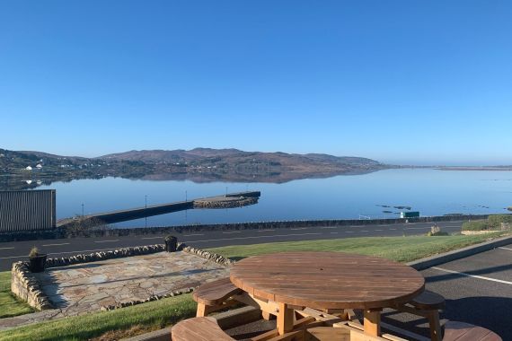 Een houten picknicktafel kijkt uit over een kalm water met een helderblauwe lucht. Op de achtergrond rijzen heuvels zachtjes op langs de horizon. Een stenen platform en goed onderhouden gras leiden naar een weg, waarachter een kalme baai de lucht weerspiegelt. Een verre pier strekt zich uit in het water. | Echt Ierland Vakanties