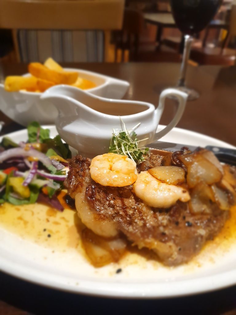 Een gegrilde steak met garnalen en ui, gegarneerd met microgreens op een wit bord. Een salade met paarse ui en komkommer. Een juskom met saus en een kom dikgesneden frieten staan op de achtergrond. Een glas rode wijn is zichtbaar op de houten tafel. | Echt Ierland Vakanties