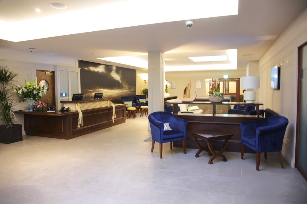 Moderne hotellobby met een receptiebalie aan de linkerkant, met een houten afwerking en een grote muurschildering erachter. Blauwe fauteuils en een ronde tafel met een lamp zijn aan de rechterkant geplaatst. Neutrale tegels bedekken de vloer en potplanten sieren de ruimte. | Echt Ierland Vakanties