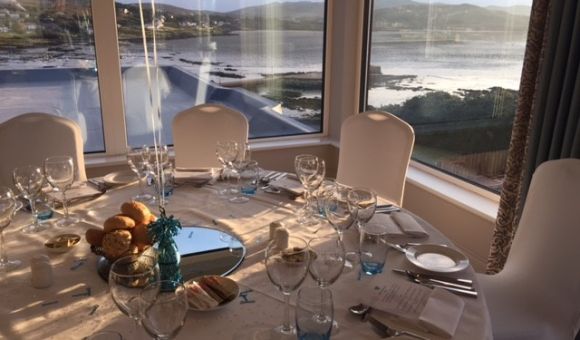 Een ronde tafel voor een formele maaltijd staat bij grote ramen met uitzicht op een serene kust. De tafel is voorzien van glaswerk, witte borden, broodjes, menukaarten en een middelpunt met een ballon. Buiten zijn kalm water en heuvels zichtbaar onder een gedeeltelijk bewolkte lucht. | Echt Ierland Vakanties