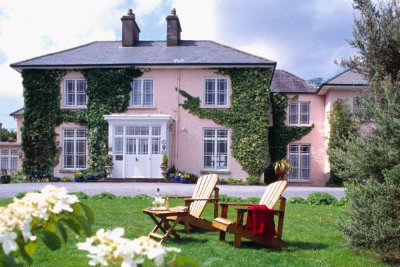 Een groot, roze huis met twee verdiepingen, met klimop die langs de muren en in de vele ramen omhoog klimt, staat achter een groen gazon. Twee houten stoelen en een tafeltje met drankjes staan op het gras, en witte bloemen staan op de voorgrond. De lucht is half bewolkt. | Echt Ierland Vakanties