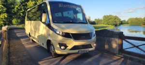 Een gele tourbus met Martours op de voorruit staat geparkeerd op een smalle brug over een rivier. De heldere, zonnige dag benadrukt groene bomen en grasvelden op de achtergrond onder een blauwe lucht met verspreide wolken. | Echt Ierland Vakanties