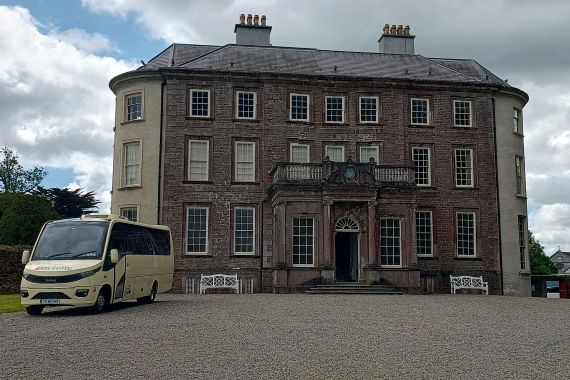 Een crèmekleurige minibus staat geparkeerd voor een groot Georgiaans herenhuis van drie verdiepingen met veel ramen, een centrale deuropening en een oprijlaan van grind. Het huis is omgeven door groene gazons, bomen en een gedeeltelijk bewolkte hemel. | Echt Ierland Vakanties