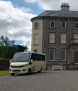 Een crèmekleurige minibus staat geparkeerd voor een groot Georgiaans herenhuis van drie verdiepingen met veel ramen, een centrale deuropening en een oprijlaan van grind. Het huis is omgeven door groene gazons, bomen en een gedeeltelijk bewolkte hemel. | Echt Ierland Vakanties