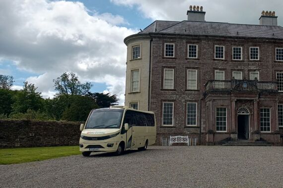 Een crèmekleurige minibus staat geparkeerd voor een groot Georgiaans herenhuis van drie verdiepingen met veel ramen, een centrale deuropening en een oprijlaan van grind. Het huis is omgeven door groene gazons, bomen en een gedeeltelijk bewolkte hemel. | Echt Ierland Vakanties