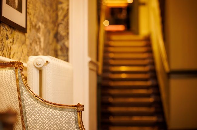 Een oude beklede stoel staat op de voorgrond bij een witte radiator, met behang met patroon op de muur. Op de achtergrond leidt een zwak verlichte trap naar boven, geaccentueerd door warm, goudkleurig licht. De focus is ondiep, waardoor de trap onscherp is. | Echt Ierland Vakanties