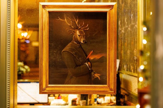 Een ingelijst schilderij toont een figuur met een hertenkop en gewei, gekleed in formele 19e-eeuwse kleding en met een wandelstok in de hand. Het portret weerspiegelt in een goudomrande spiegel, met warme sfeerverlichting en vage decoratieve lampen op de achtergrond. | Echt Ierland Vakanties