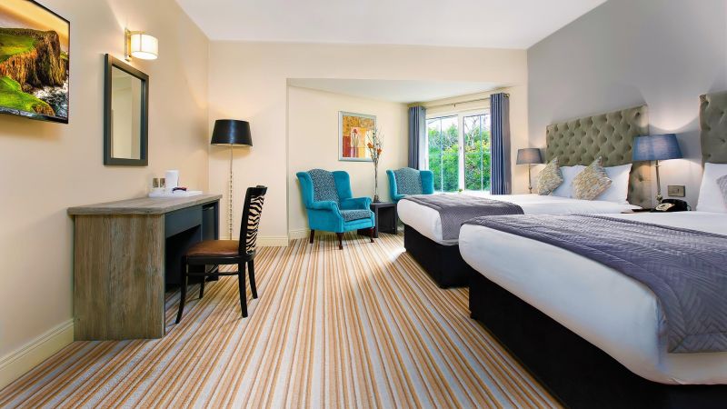 Een moderne Hillyard House Hotel kamer in Castlewellan met twee tweepersoonsbedden, groenblauwe bedlampjes, een gestreept tapijt, een houten bureau en stoel, een grote spiegel en groenblauwe fauteuils bij het raam. Kunst siert de crèmekleurige en grijze muren terwijl het daglicht naar binnen stroomt. | Echt Ierland Vakanties