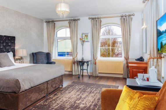 Een ruime, zonovergoten kamer in het Hillyard House Hotel in Castlewellan heeft een groot bed met grijs beddengoed, een fauteuil, flatscreentelevisie, een tafeltje met bloemen en twee boogramen. Warme verlichting en een moderne inrichting creëren een gezellige, uitnodigende sfeer. | Echt Ierland Vakanties