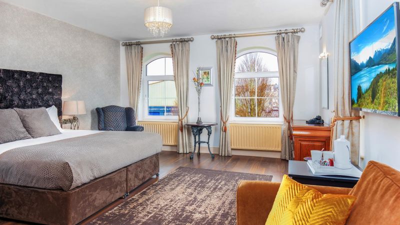 Een ruime, zonovergoten kamer in het Hillyard House Hotel in Castlewellan heeft een groot bed met grijs beddengoed, een fauteuil, flatscreentelevisie, een tafeltje met bloemen en twee boogramen. Warme verlichting en een moderne inrichting creëren een gezellige, uitnodigende sfeer. | Echt Ierland Vakanties