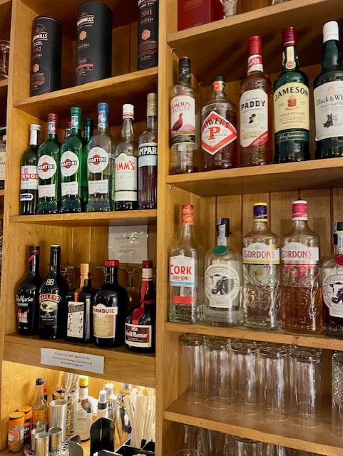 Op houten planken staan verschillende flessen drank, waaronder Gordon's gin, Jameson whiskey, Aperol, Campari en Baileys. Daaronder staat glaswerk netjes gerangschikt. Andere whiskey- en likeurmerken zijn zichtbaar, met cocktailbenodigdheden op een lagere plank. | Echt Ierland Vakanties