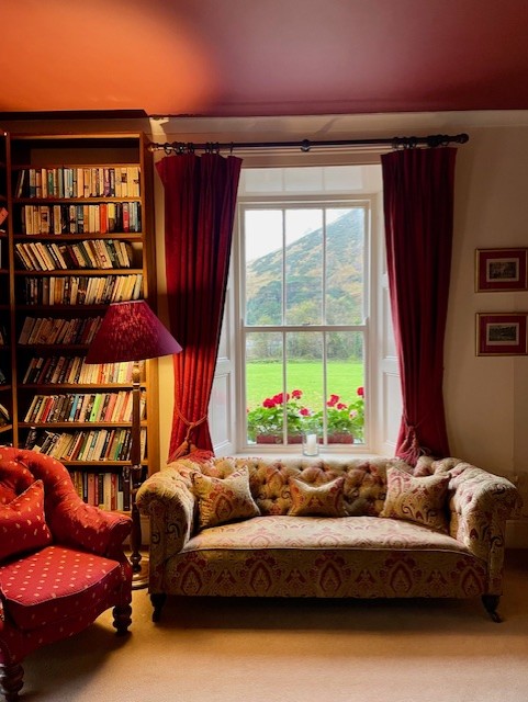 Een gezellige woonkamer heeft een bank met patroon en een rode fauteuil naast een hoge boekenkast gevuld met boeken. Een raam met rode gordijnen biedt uitzicht op een groen gazon, rode bloemen en heuvels buiten. Warme verlichting creëert een uitnodigende sfeer. | Echt Ierland Vakanties