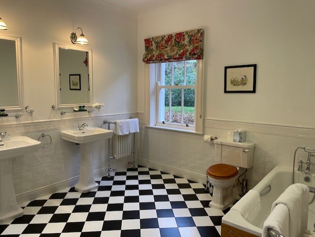 Een ruime badkamer met een zwart-wit geblokte vloer, twee wastafels met spiegels en wandlampen, een raam met een bloemengordijn, een houten toilet, een bad met een witte handdoek en wit betegelde muren versierd met ingelijste kunst. | Echt Ierland Vakanties