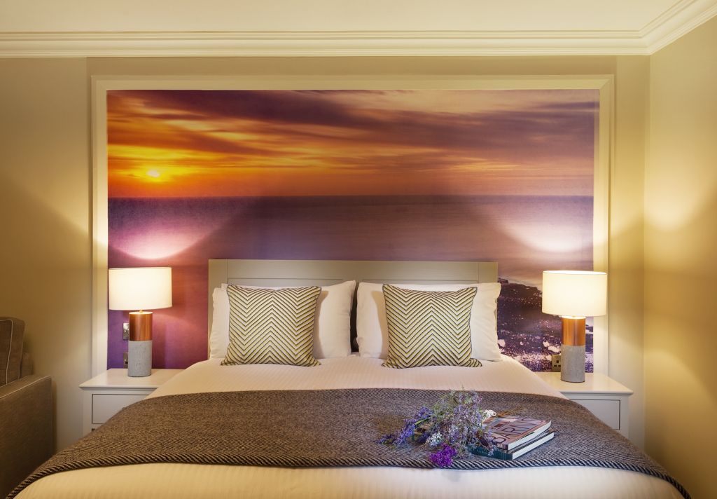 Een gezellige hotelslaapkamer met een bed tegen een grote muurschildering van een zonsondergang boven het water. Het bed heeft twee kussens met een zigzagpatroon, een grijs kleed en nachtkastjes met aan elke kant een lamp. Een tijdschrift en een takje bloemen liggen op het bed. | Echt Ierland Vakanties