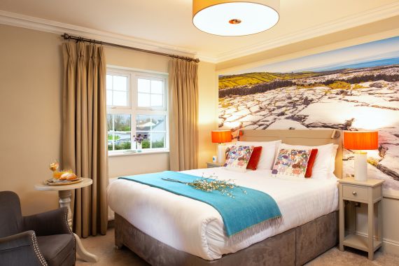 Een gezellige slaapkamer met een groot bed, wit linnengoed, een blauw kleed en kleurrijke kussens. Twee lampen op bijzettafeltjes flankeren het bed. Een landschap muurschildering siert de muur. Een raam met beige gordijnen laat licht binnen. Een klein tafeltje met een drankje en gebakje staat bij een grijze leunstoel. | Echt Ierland Vakanties