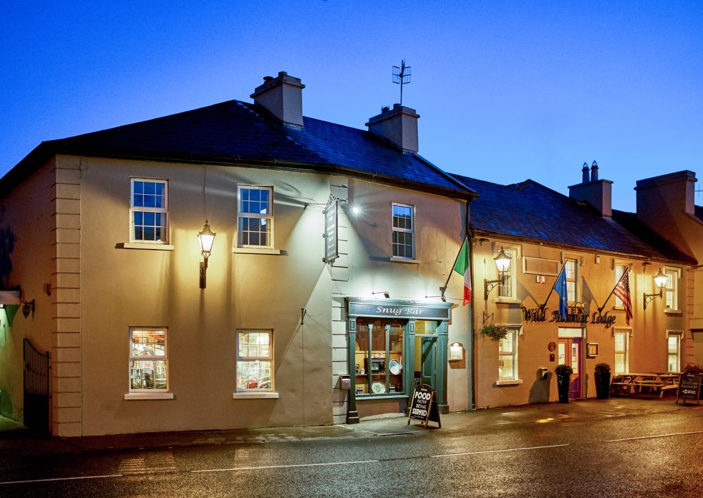Een warm verlichte Ierse pub en winkel in de schemering, met crèmekleurige muren, schoorstenen en hangende lantaarns. Buiten hangt een Ierse vlag en bij de deur hangen schoolbordbordjes. De natte straat weerspiegelt de lichten onder een diepblauwe avondhemel. | Echt Ierland Vakanties