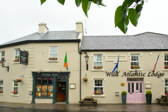 Een crèmekleurig gebouw met de naam "Wild Atlantic Lodge", roze dubbele deuren, bloembakken op de vensterbanken, Ierse en andere vlaggen en een uithangbord voor Snug Bar. Buiten aan een natte straat staan banken onder bladerrijke takken. | Echt Ierland Vakanties