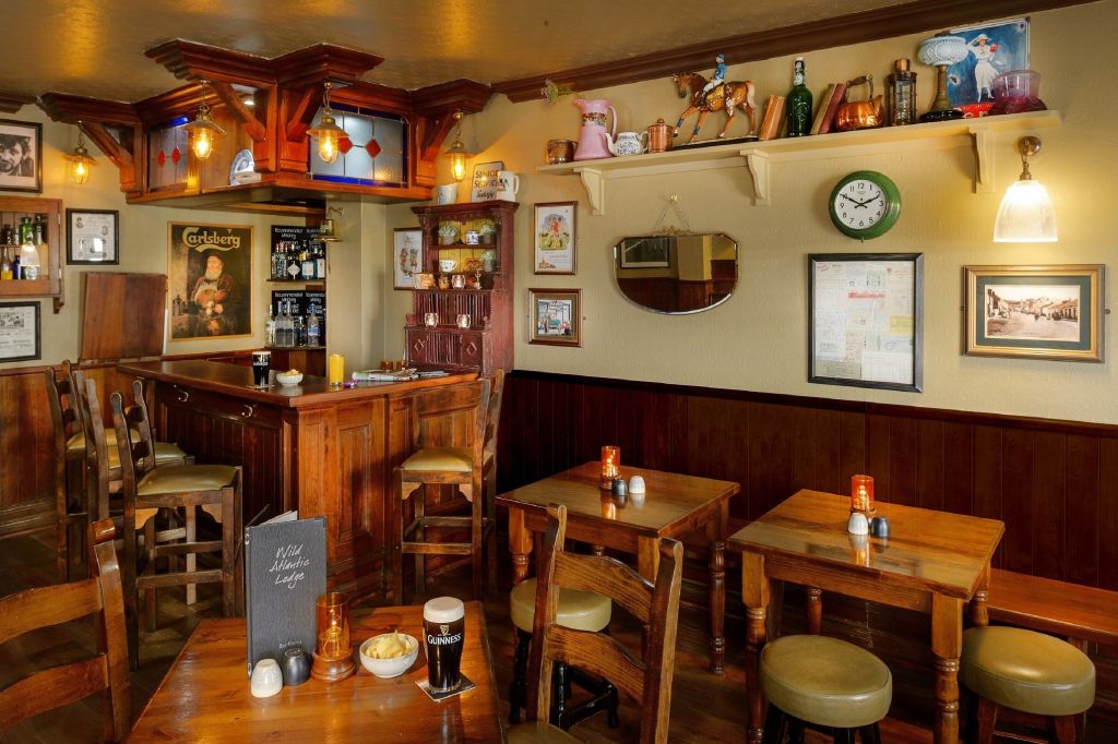 Gezellig pubinterieur met houten tafels en stoelen, zachtgele verlichting en een klein bargedeelte. Op planken staat eigenzinnige decoratie zoals theepotten en modelhonden. Kaarsen, hapjes en drankjes staan op tafels; ingelijste kunst en een klok hangen aan beige muren. | Echt Ierland Vakanties