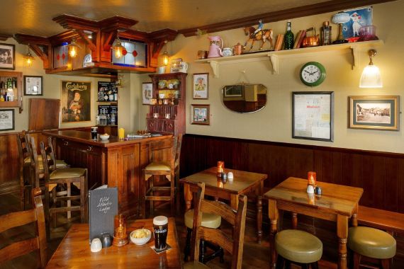 Gezellig pubinterieur met houten tafels en stoelen, zachtgele verlichting en een klein bargedeelte. Op planken staat eigenzinnige decoratie zoals theepotten en modelhonden. Kaarsen, hapjes en drankjes staan op tafels; ingelijste kunst en een klok hangen aan beige muren. | Echt Ierland Vakanties