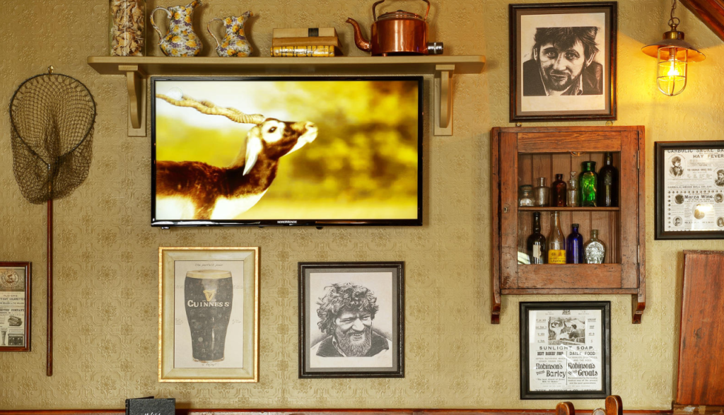 Een warm verlichte muur versierd met ingelijste foto's, vintage flessen, een visnet en een tv met een antilope. Het decor bestaat uit een Guinness-afdruk, sepiaportretten, rustieke containers en een wandlamp, die een gezellige, eclectische sfeer creëren. | Echt Ierland Vakanties