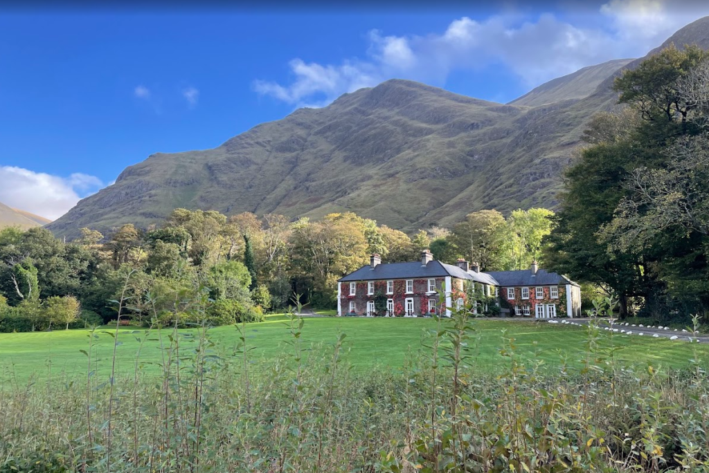 Een groot bakstenen landhuis met witte sierranden en meerdere schoorstenen staat op een ruim groen gazon, omringd door bomen. Achter het huis rijzen hoge ruige bergen steil omhoog onder een helderblauwe hemel met verspreide wolken. | Echt Ierland Vakanties