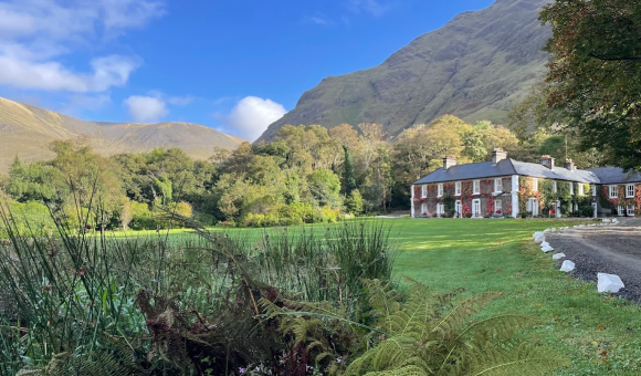 Een groot bakstenen huis met wit omrande ramen staat op een weelderig groen gazon, omringd door bomen. Hoge, steile heuvels rijzen op in de achtergrond onder een blauwe lucht met verspreide wolken. Varens en planten verschijnen op de voorgrond bij een gebogen oprijlaan van grind. | Echt Ierland Vakanties