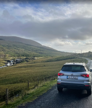 Een zilverkleurige SUV met Ierse nummerborden staat geparkeerd op een smalle, kronkelige weg langs een weelderig groene vallei. Bergen en heuvels strekken zich in de verte uit onder een gedeeltelijk bewolkte hemel. Links in het landschap stroomt een riviertje met stroomversnellingen. | Echt Ierland Vakanties