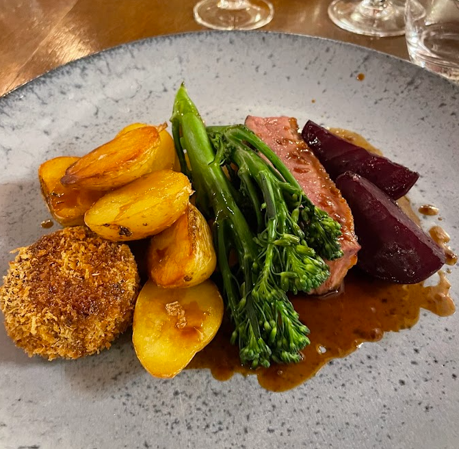 Een gastronomisch gerecht met geroosterde aardappelen, malse broccoli, een plakje roze medium gegaard vlees met bruine saus, twee stukjes geglaceerde rode biet en een goudbruin gepaneerde kroket, geserveerd op een grijs keramisch bord. | Echt Ierland Vakanties