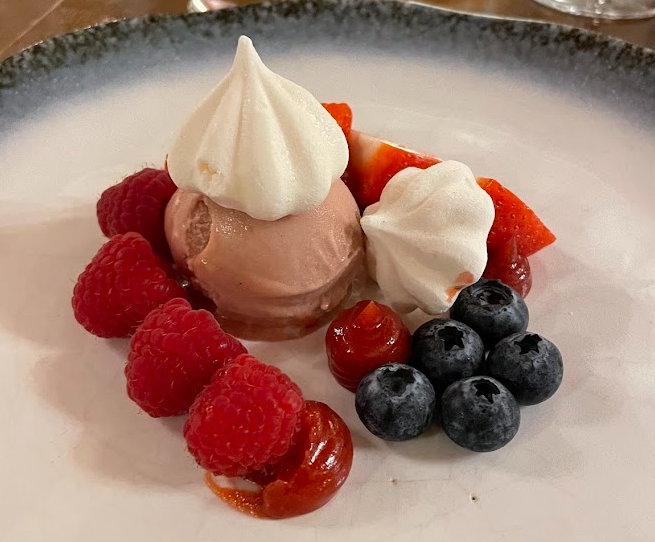 Een dessertbord met een bolletje roze roomijs met daarop twee witte meringues, omringd door verse frambozen, bosbessen, plakjes aardbeien en kleine toefjes rode saus, geserveerd op een wit bord met een donkere rand. | Echt Ierland Vakanties