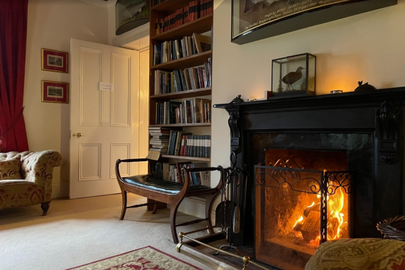Een gezellige woonkamer met een brandende open haard, een sierlijke zwarte schoorsteenmantel en haardgereedschap ervoor. Boekenplanken vol boeken staan naast een open witte deur. Twee fauteuils met patroon, een houten bank, rode gordijnen, ingelijste kunst en warme verlichting maken het uitnodigende tafereel compleet. | Echt Ierland Vakanties
