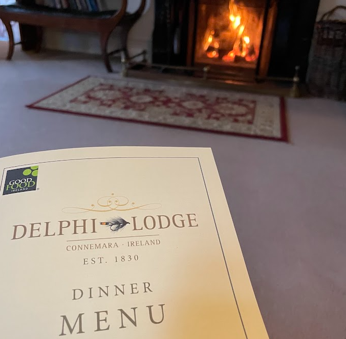 Een close-up van een Delphi Lodge Dinner Menu met Connemara, Ireland Est. 1830 erop gedrukt, gehouden in een gezellige woonkamer. Op de achtergrond een brandende open haard, een tapijt met patroon en een deel van een houten stoel. | Echt Ierland Vakanties