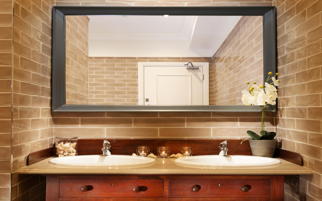 Een moderne badkamer heeft beige metro tegelwanden, een grote rechthoekige spiegel, een houten badmeubel met twee witte wastafels, zilveren kranen, kaarsen, een glazen pot met schelpen en een witte orchidee in pot op het aanrecht. Een witte deur is zichtbaar in de spiegel. | Echt Ierland Vakanties