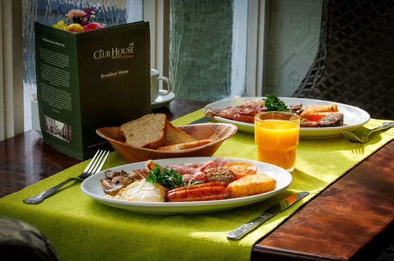 Twee borden met een volledig Engels ontbijt met eieren, bacon, worst, hash browns, gegrilde tomaat, champignons en bonen staan op een groene placemat. Er staat een glas sinaasappelsap, een mandje brood, bestek en een groen ontbijtmenu op de donkere houten tafel. | Echt Ierland Vakanties