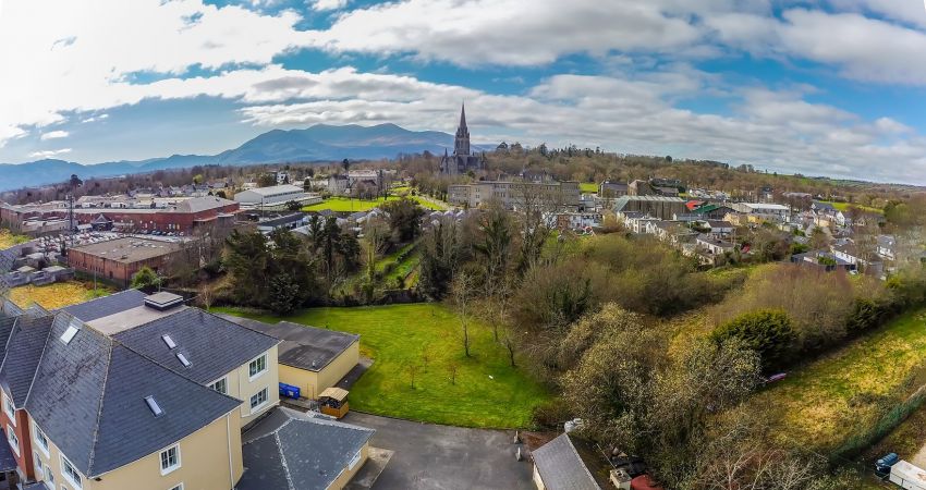 Luchtfoto van Killarney met verspreide huizen, een kerk met een hoge toren en veel groen. Het Brook Lodge Hotel nestelt zich tussen stedelijke en natuurlijke elementen, terwijl bergen en wolken het vredige tafereel vullen onder een gedeeltelijk bewolkte blauwe hemel. | Echt Ierland Vakanties