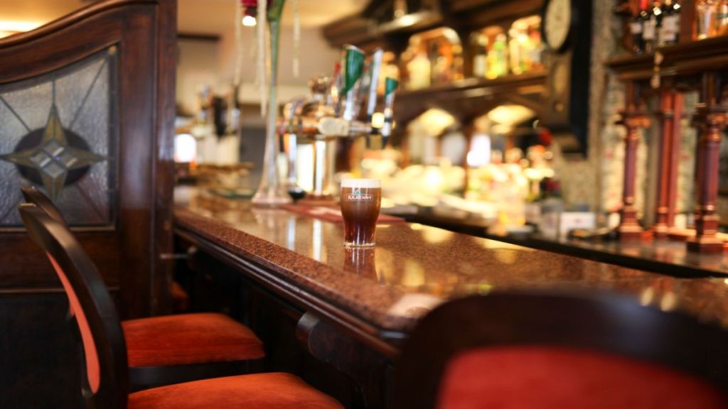 Een glas donker bier staat op een gepolijste houten bar in een gezellige pub met rood gestoffeerde stoelen, sierlijk houtwerk, bierkranen en planken vol flesjes en glaswerk wazig op de achtergrond. | Echt Ierland Vakanties