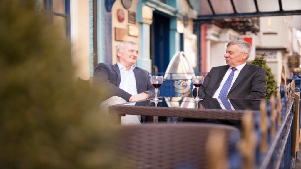 Twee oudere mannen in pak zitten aan een cafétafel buiten, glimlachen en praten. Ze hebben elk een glas rode wijn voor zich. De omgeving wordt gekenmerkt door blauwe balustrades, groen en kleurrijke gevels op de achtergrond. | Echt Ierland Vakanties